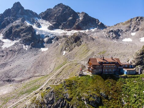 A mountain cabin surrounded by impressive mountains and glaciers. The landscape is green and offers a clear view of the surrounding peaks.