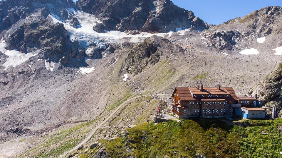 A mountain cabin surrounded by impressive mountains and glaciers. The landscape is green and offers a clear view of the surrounding peaks.