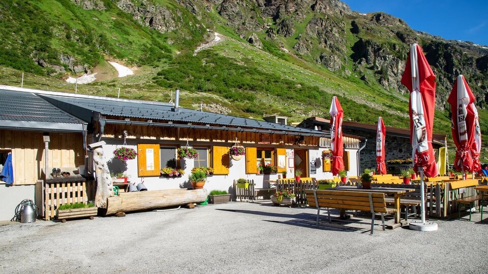 A cozy alpine hut with colorful flowers and red umbrellas. In the background, green mountains can be seen.
