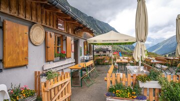 A cozy mountain cabin with sun umbrellas and tables outside. Majestic mountains and a cloudy sky can be seen in the background.