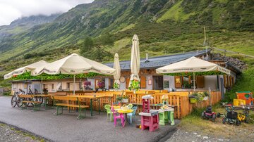 A cozy mountain cabin with tables and sun umbrellas outside. The surroundings are green and mountainous, perfect for excursions into nature.