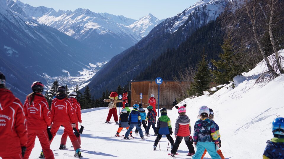 A group of children and ski instructors on a ski slope in the mountains. The surroundings are snowy and it is sunny.