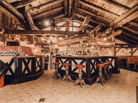 A cozy bar in a rustic ambiance with wooden ceilings and beams. Red bar stools surround the central counter.