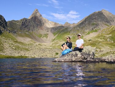 Zwei Personen sitzen auf einem Felsen am Ufer eines Sees in den Bergen. Die Landschaft ist majestätisch mit hohen Gipfeln und klarem Himmel.