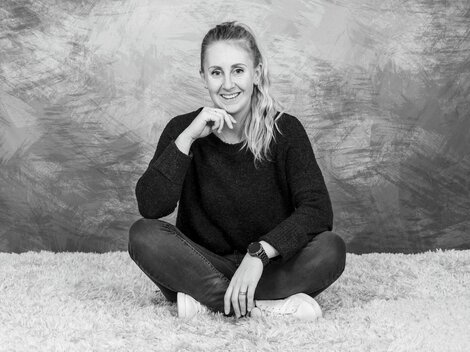 A young woman is smiling while sitting cross-legged on a soft carpet. She is wearing a dark sweater and jeans. | © Simone Drexel Photography