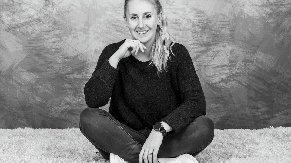 A young woman is smiling while sitting cross-legged on a soft carpet. She is wearing a dark sweater and jeans. | © Simone Drexel Photography