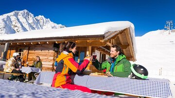 Ein Paar genießt Getränke in einem Schneegebiet vor einer Berghütte. Im Hintergrund sind schneebedeckte Berge und weitere Besucher zu sehen.