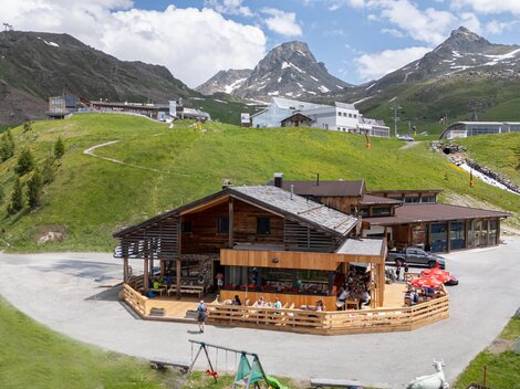 A modern wooden house in the mountains, surrounded by green meadows and rugged peaks. The sky is blue with some white clouds.