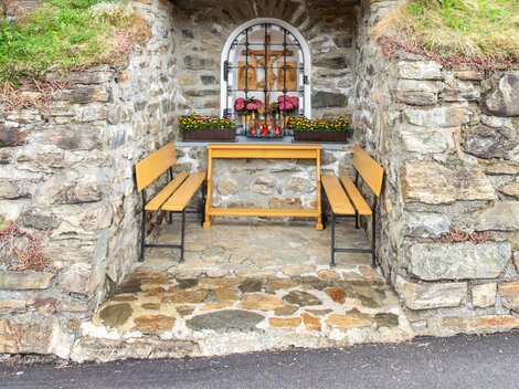 A small, rustic chapel made of stone and wood stands in a green environment. In front of the chapel, two benches and flowers can be seen. | © Bernhard Gruber – Photography