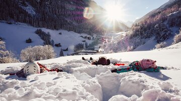 Children lie in the snow, surrounded by a picturesque winter landscape. The sun shines brightly over the snow-covered hills.