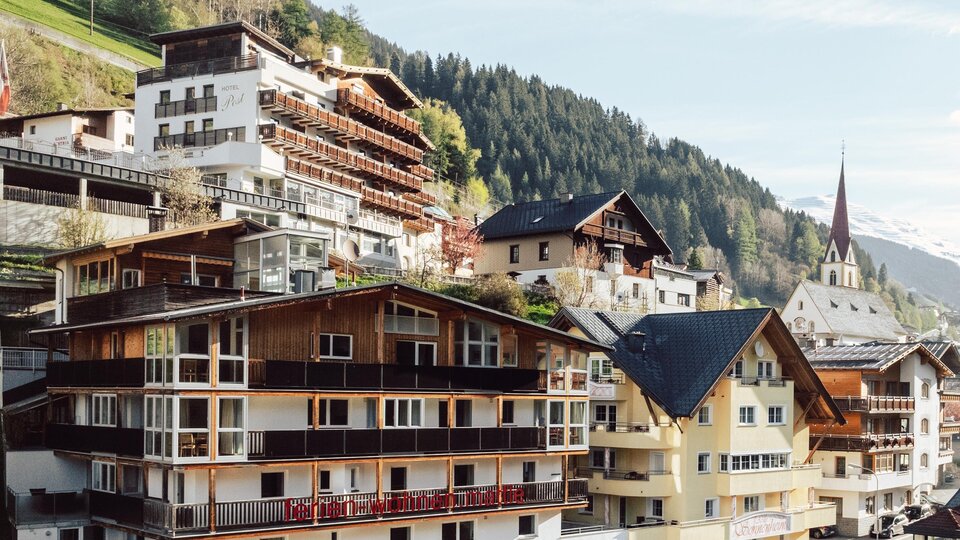 A picturesque mountain landscape with several modern chalets and traditional buildings. The houses are situated on a slope, surrounded by forests and mountains.