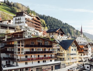 A picturesque mountain landscape with several modern chalets and traditional buildings. The houses are situated on a slope, surrounded by forests and mountains.