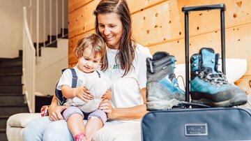 A mother sits with her child on a sofa in a modern room. Next to them are hiking shoes and a suitcase.