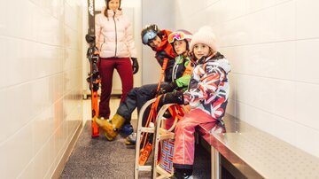 A skier is standing with skis in a corridor, while two children are sitting on a bench. The girl is wearing colorful ski clothing and a pink hat, and the boy is wearing a helmet.