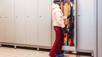 A person with ski equipment stands in front of a cabinet in a ski room. Skis and poles can be seen in the cabinet.