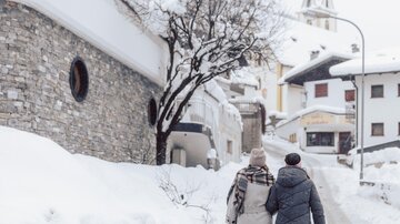 Two people are walking on a snow-covered path in a quiet village. A church tower is visible in the background.