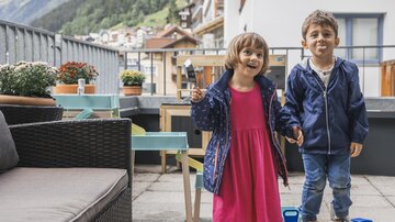 Two cheerful children are standing on a balcony. They are wearing colorful clothes and holding toys in their hands.