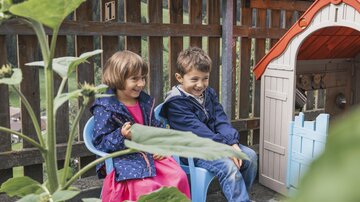 Two children are sitting on blue chairs and laughing. Behind them, there is a small playhouse in front of a wooden slat wall.