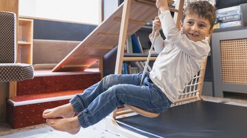 A cheerful boy is swinging on a wooden swing in a child-friendly room. In the background, a slide and bookshelves can be seen.