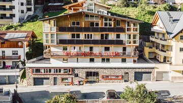 A modern multi-story building in a small town. The facade is made of wood and stone, surrounded by green hills and other houses.