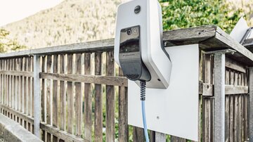 An electric charging station is mounted on a wooden railing. In the background, trees and mountains are visible.