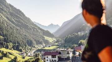 A person looks at a picturesque valley with mountains in the background. The landscape is green and the sun shines through the valleys.