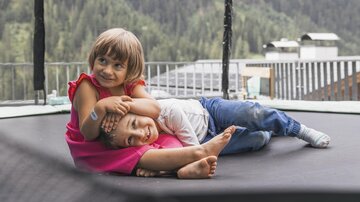 Two children are lying relaxed on a trampoline. The girl is hugging the boy while they have a beautiful view of the surrounding landscape.